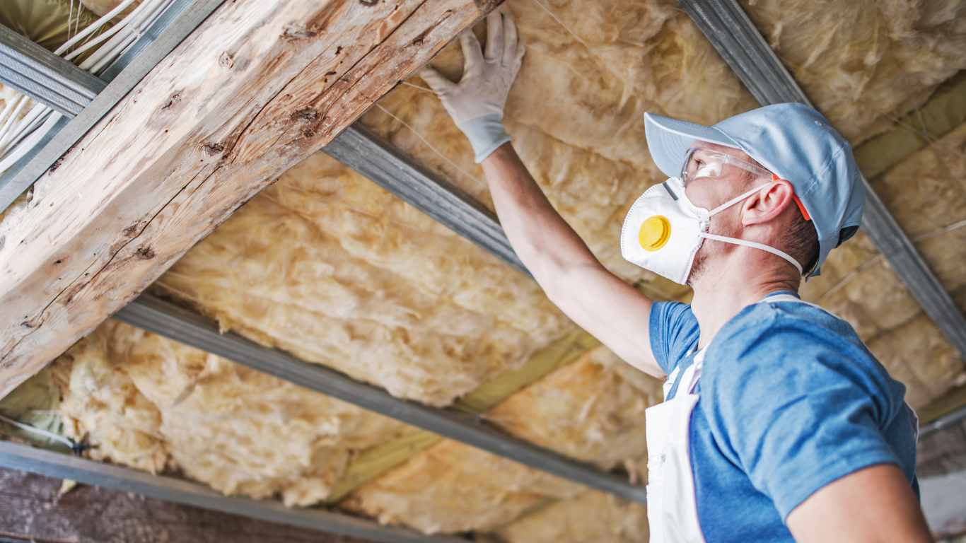 Man installing insulation in attic, wearing mask, gloves, and protective gear.