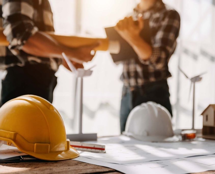 Two engineers reviewing blueprints at a table with hard hats and wind turbine models.