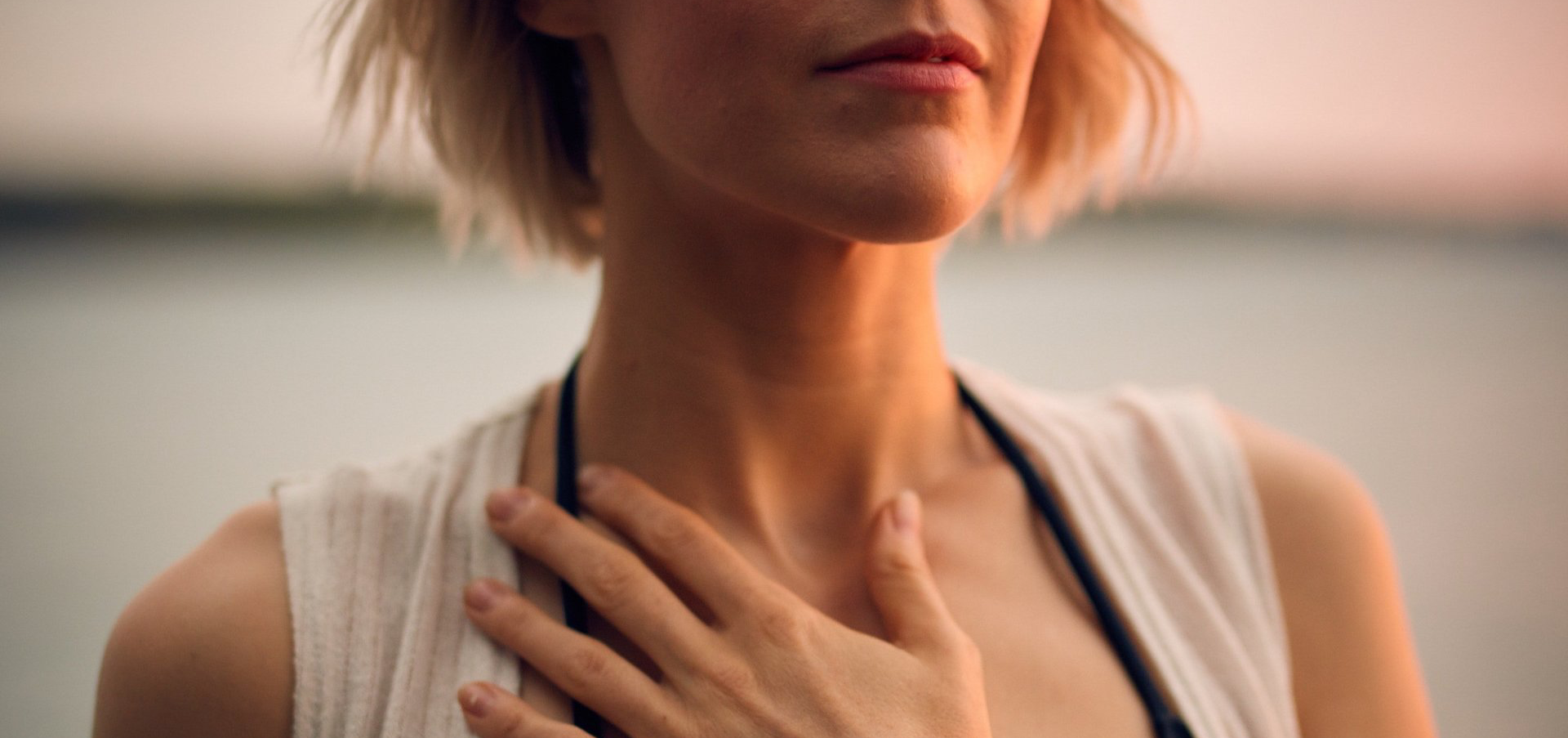 Woman with short blonde hair, hand on chest, standing near water. Soft light, relaxed expression.