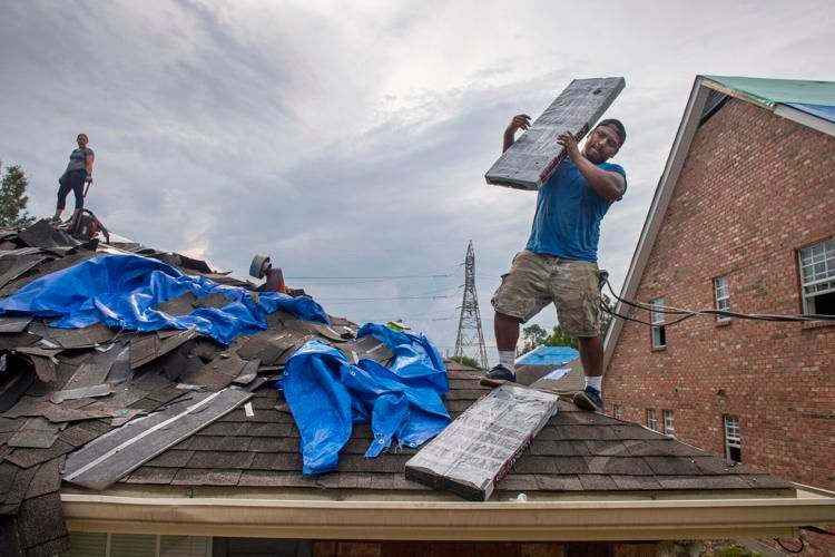 Roofers replacing shingles on a house after a storm. Workers in blue and grey clothing on a rooftop, with a cloudy sky.