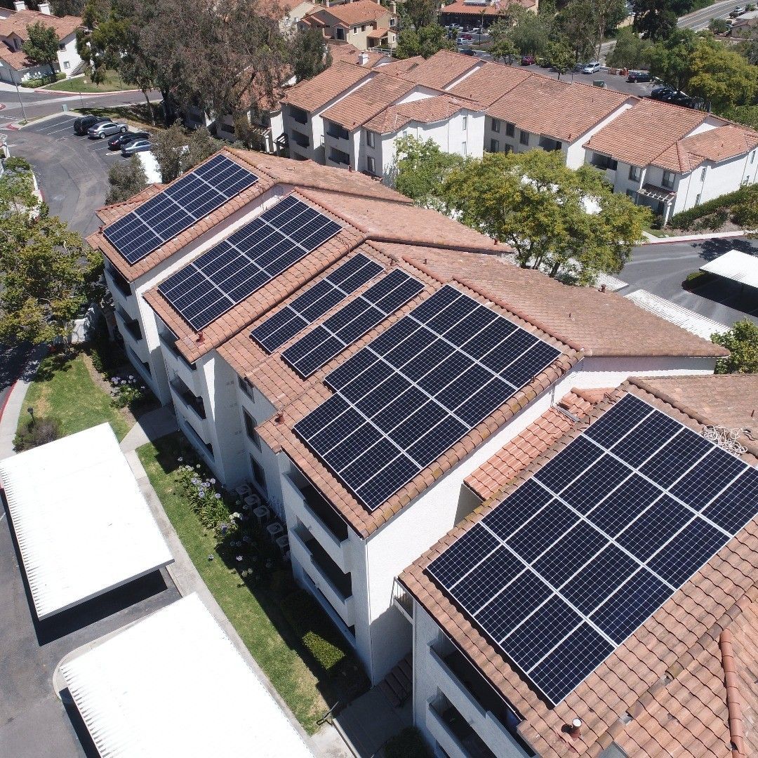 Solar panels on the red-tiled roofs of a multi-story apartment building. Green grass and trees surround it.