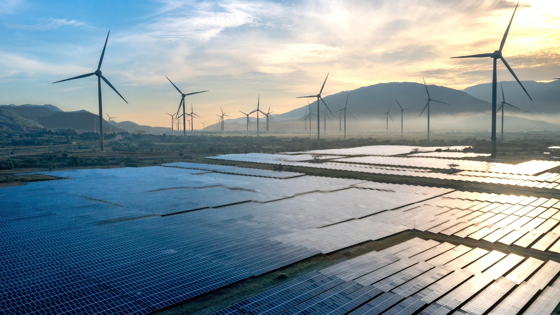 Solar panels and wind turbines generate power near mountains at sunset.