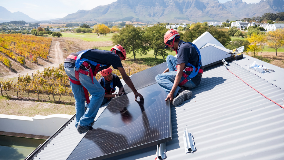Workers in safety harnesses installing solar panels on a rooftop with a mountain backdrop.