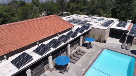 Aerial view of building with solar panels and pool. Blue umbrellas and lounge chairs.
