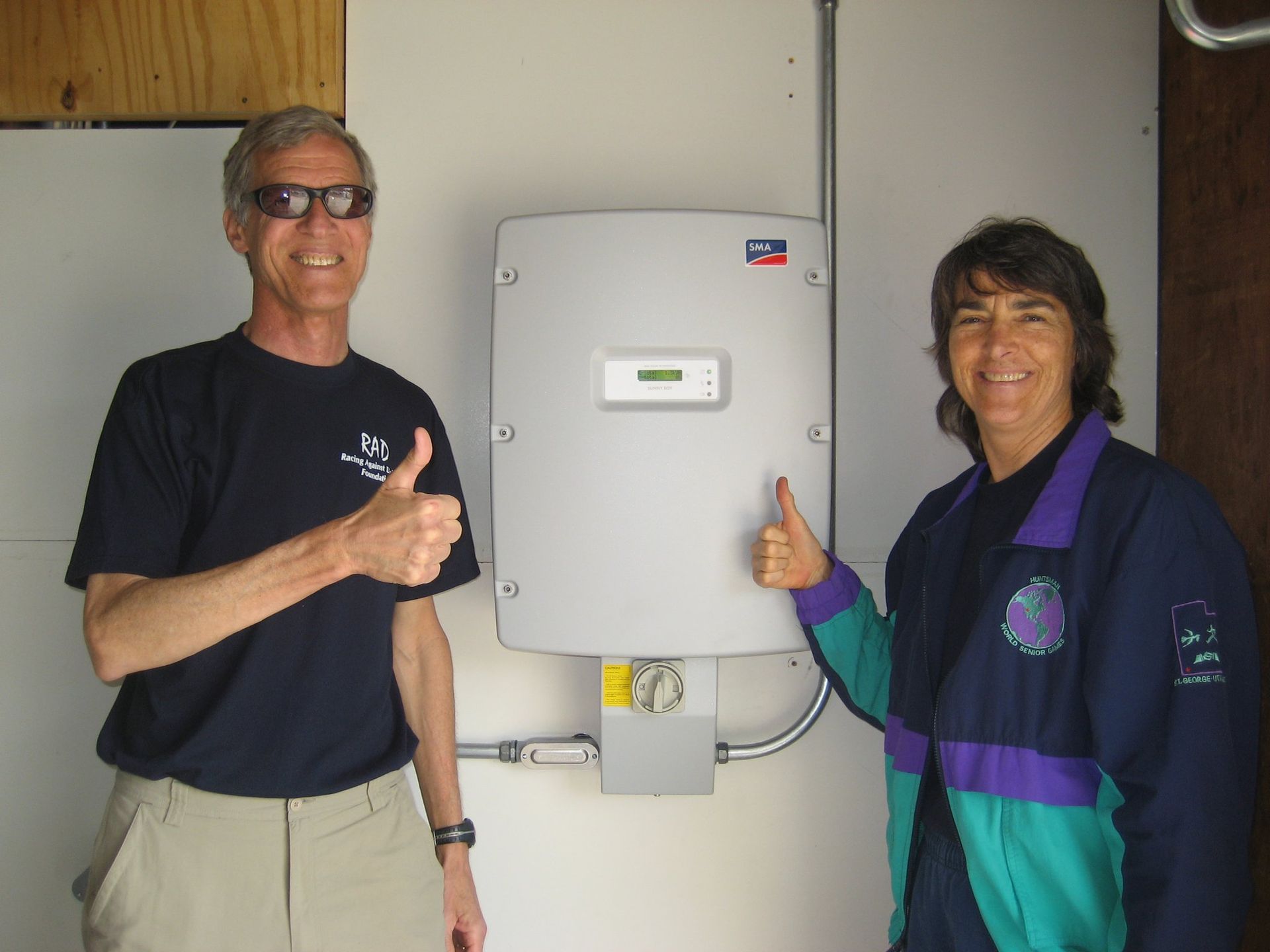 Two people give thumbs up next to a solar panel inverter on a wall.