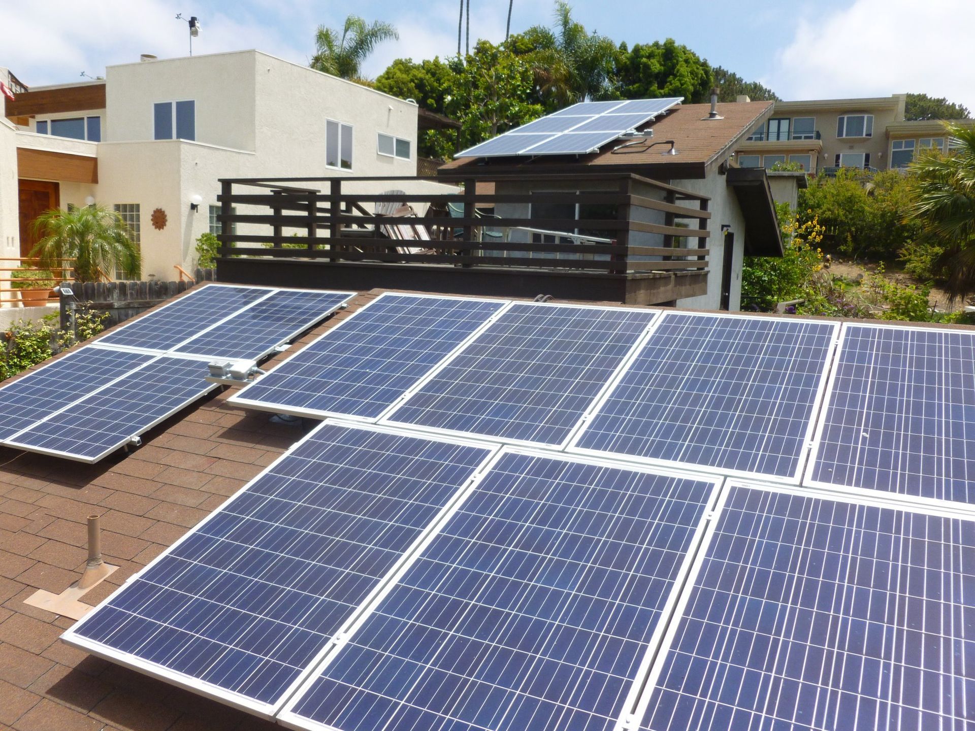 Solar panels on a brown-shingled roof in front of a white building with a deck.