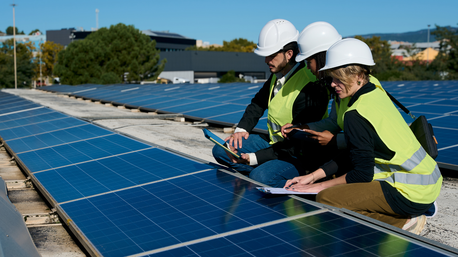 Three people in safety vests and hard hats inspecting solar panels on a rooftop.