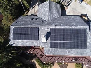 Aerial view of a house with solar panels installed on a gray shingle roof. A deck and palm trees are visible.