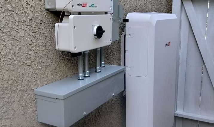 Solar panel equipment mounted on a stucco wall: white boxes, grey conduit and a large white battery.