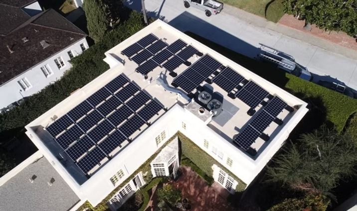 Rooftop with solar panels on a multi-sectioned white house surrounded by greenery.