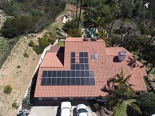 Aerial view of a home with solar panels on a red tile roof, cars in the driveway, and hillside vegetation.