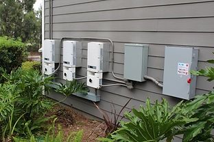 Solar panel equipment mounted on a gray house exterior, next to plants.