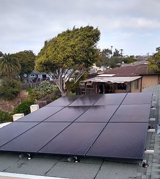 Solar panels installed on a rooftop. Houses and a tree are visible in the background, overcast sky.