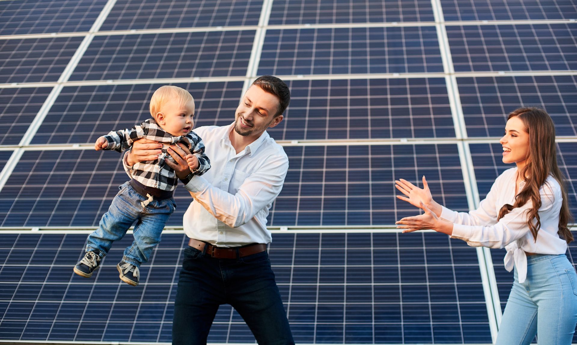 Family standing in front of solar panels, smiling and reaching toward each other outdoors.