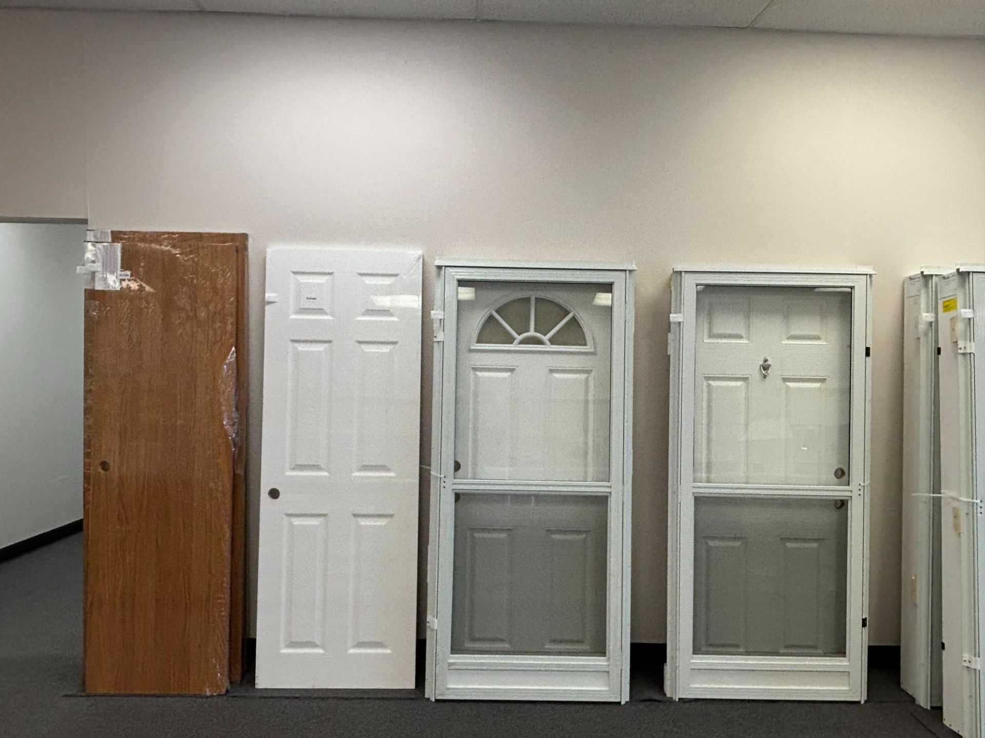 Five doors of various styles stand against a neutral wall; one is wrapped in brown paper.