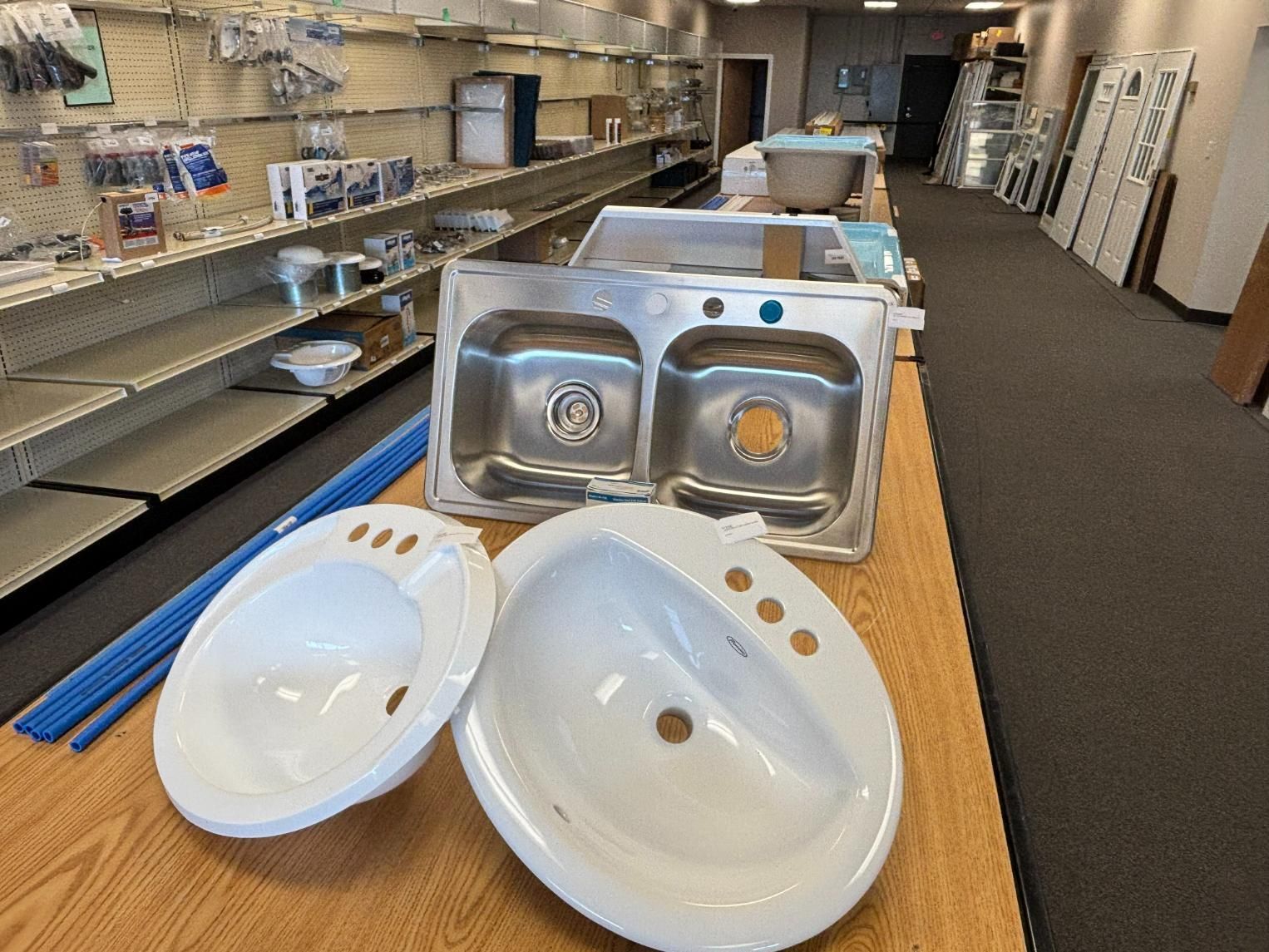 A hardware store display: stainless steel double sink and two white sinks on a table. Shelves of hardware in the background.