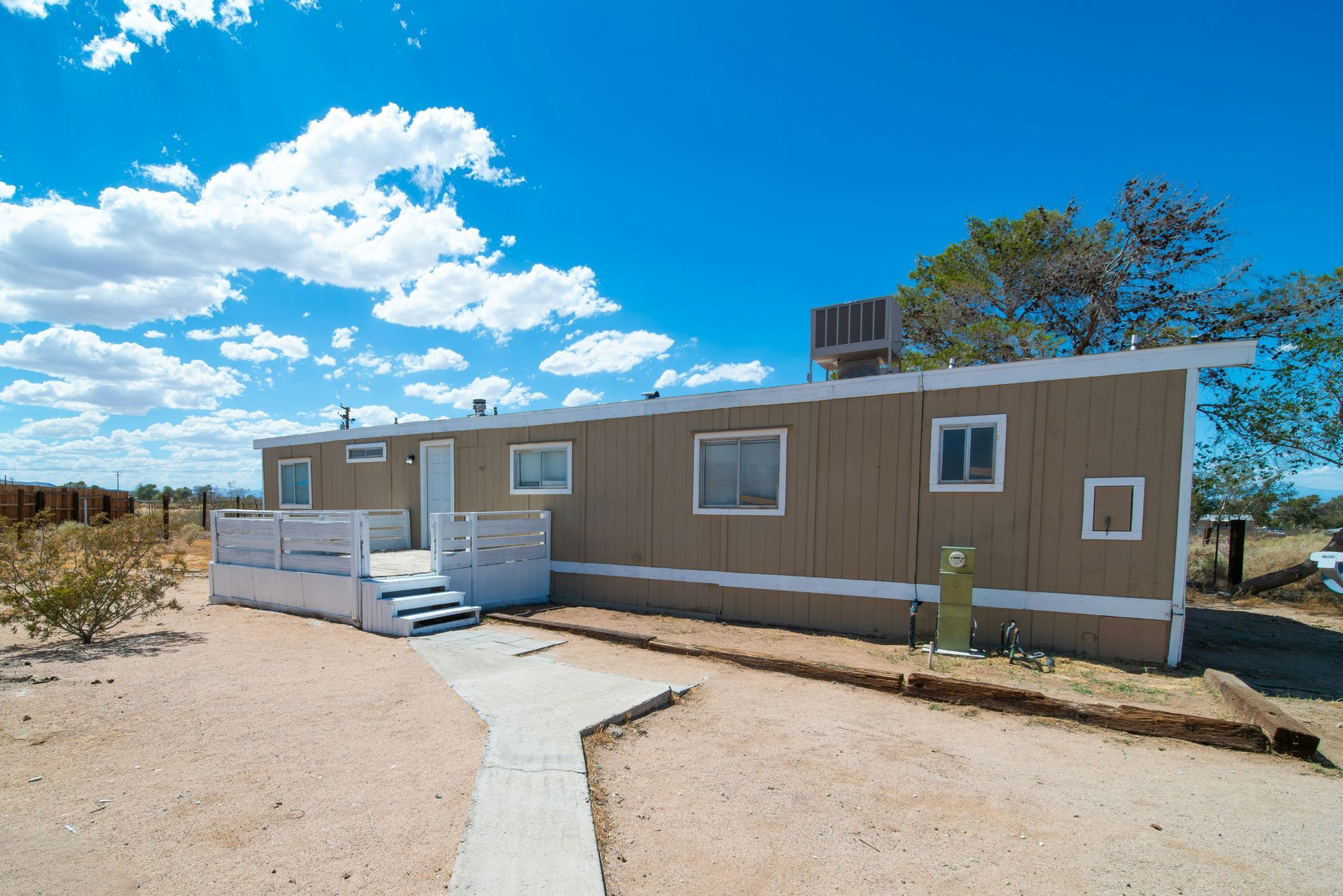 Tan mobile home on dry, sandy land under a bright blue sky with scattered clouds.