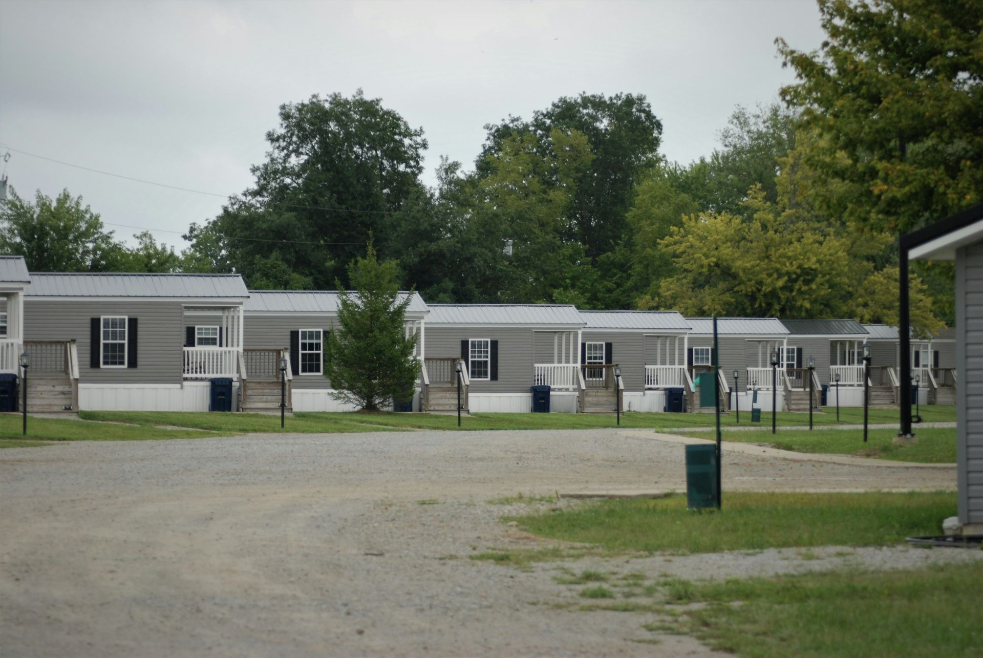 Row of gray mobile homes in a gravel lot, with trees in the background under an overcast sky.