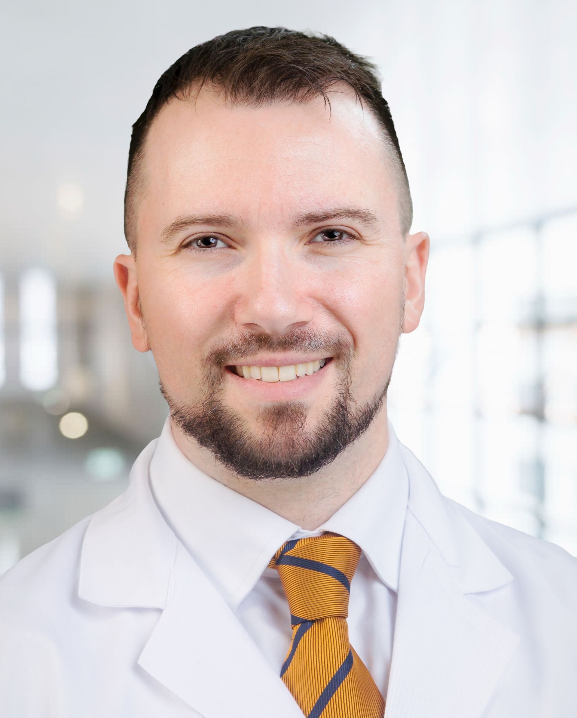 Smiling doctor in a white coat and yellow tie, posed in a bright clinic setting.