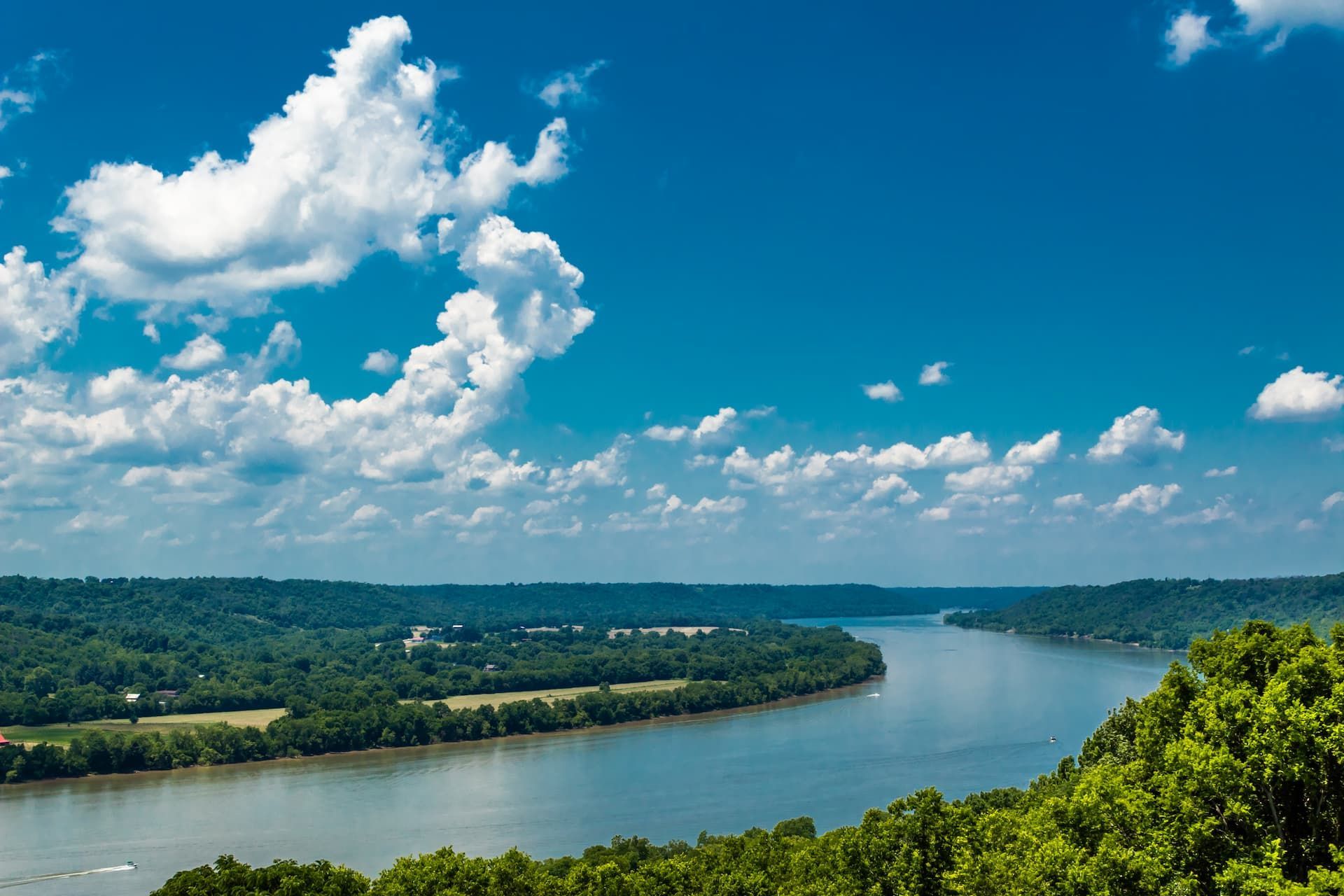 River winding through lush green hills under a bright blue sky dotted with puffy white clouds.