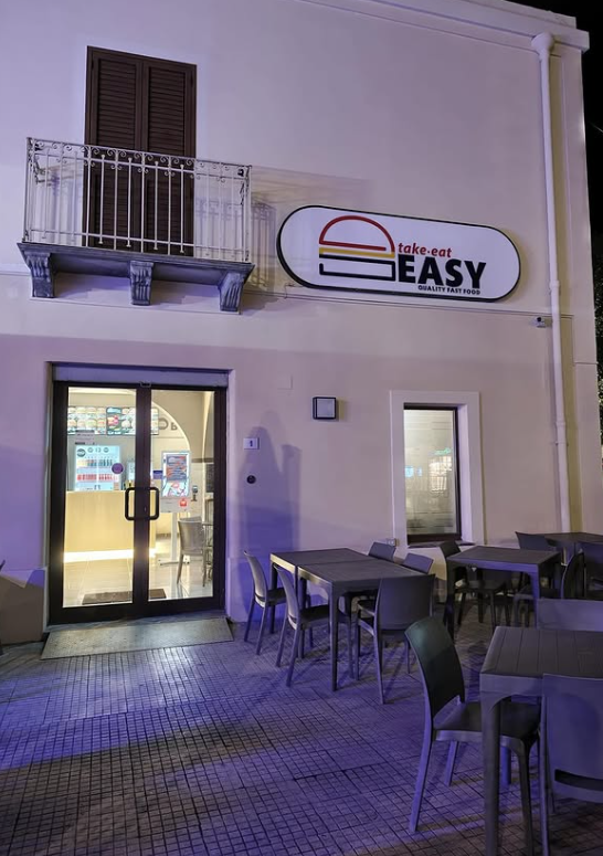 Piccola caffetteria illuminata di notte con tavolini all'aperto e balcone, insegna con la scritta