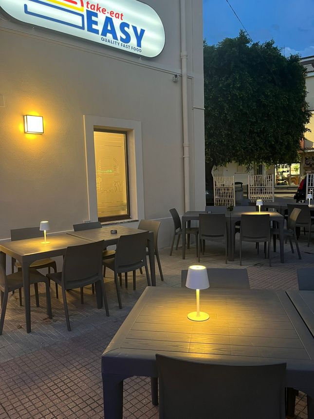 Al crepuscolo, accanto a un edificio color crema, si possono trovare tavolini all'aperto illuminati e un'insegna luminosa, con tavolini e insegne al neon.