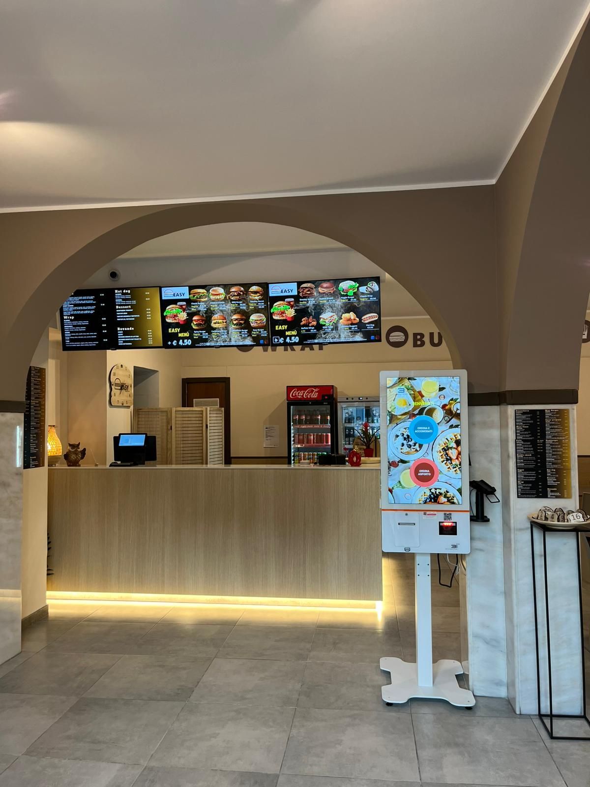 Bancone di una caffetteria con menù digitali, illuminazione calda e insegna autoportante in un moderno centro commerciale al coperto.