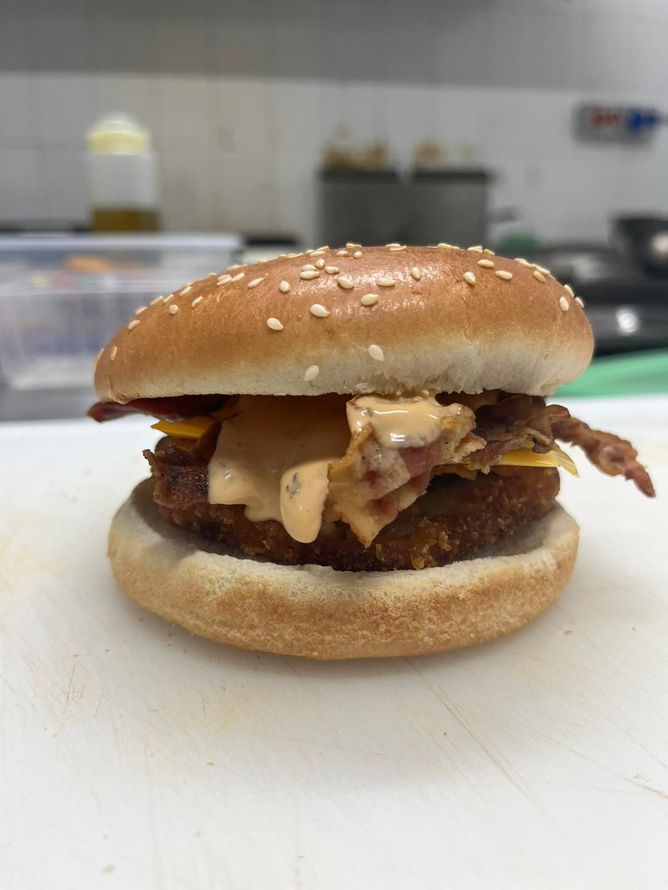 Hamburger con semi di sesamo, formaggio fuso e pancetta, servito su un tagliere bianco.