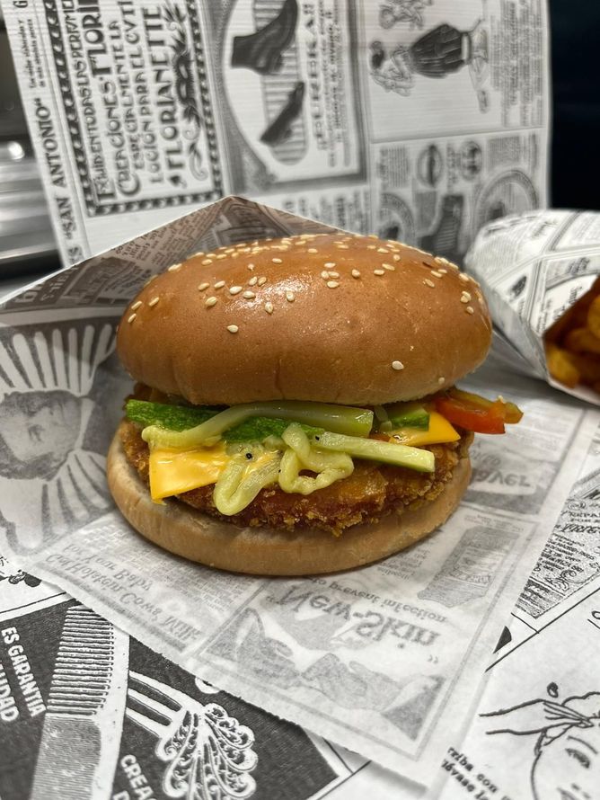 Hamburger al sesamo con lattuga, formaggio e patatine fritte su un vassoio foderato di giornale, con il sacchetto delle patatine sullo sfondo.