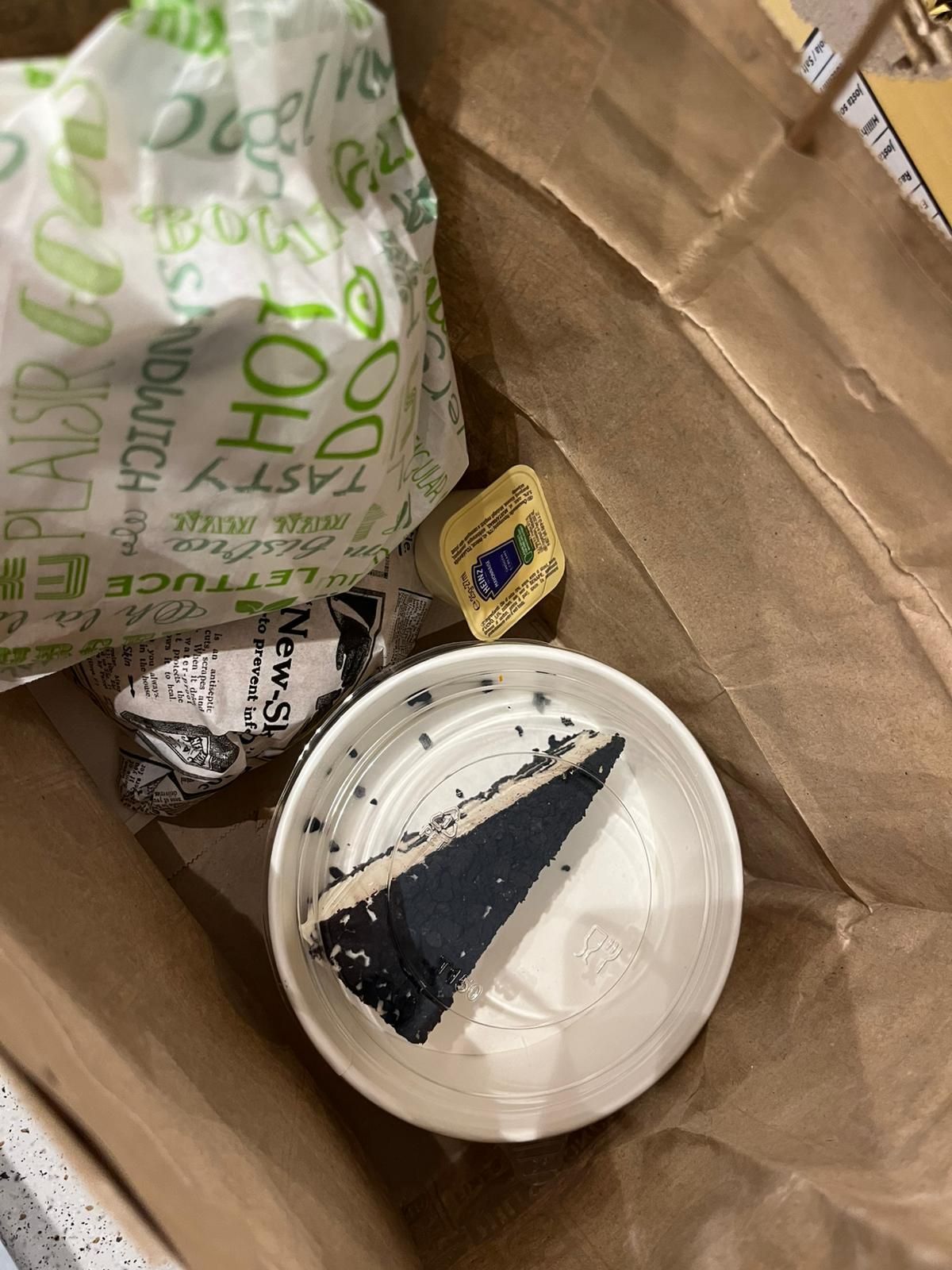 Sacchetto di carta con contenitori per cibo da asporto, una fetta di torta al cioccolato e una piccola bustina di condimento all'interno.