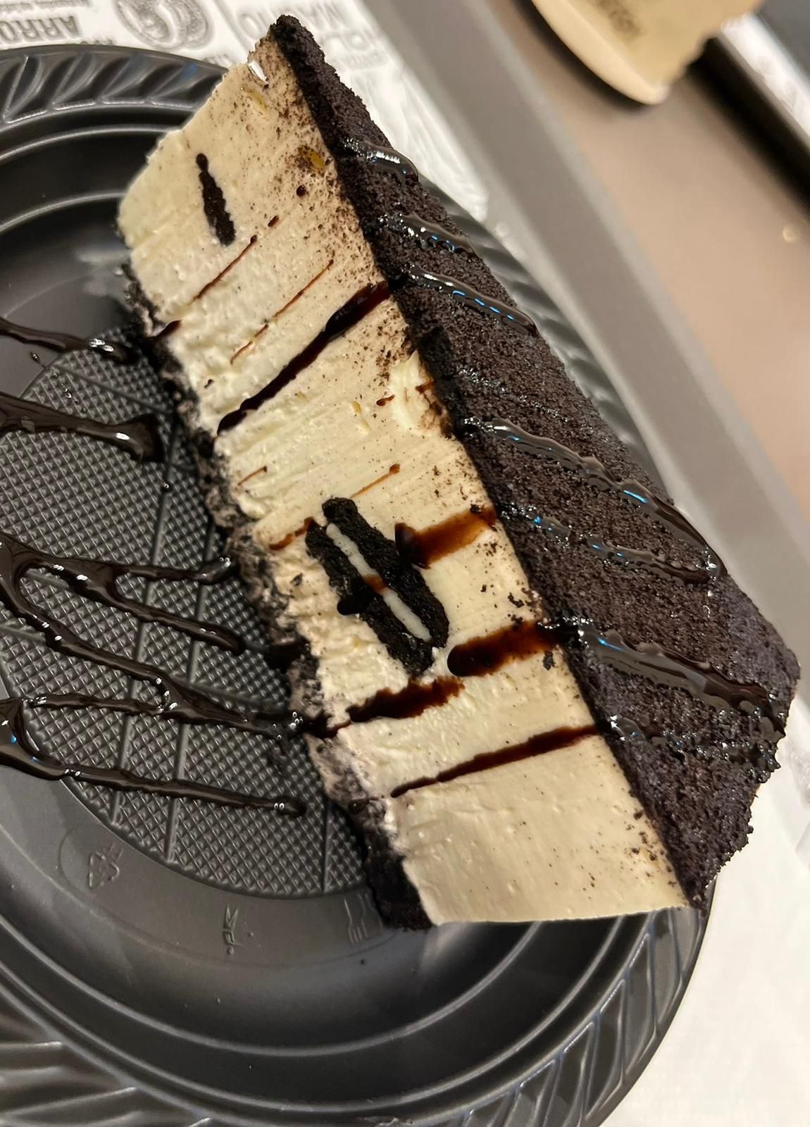 Una fetta di cheesecake con glassa al cioccolato su un piatto di plastica nero.