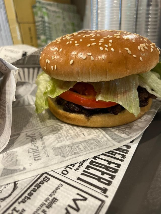 Hamburger con semi di sesamo, lattuga e pomodoro, avvolto in carta e appoggiato su un bancone.