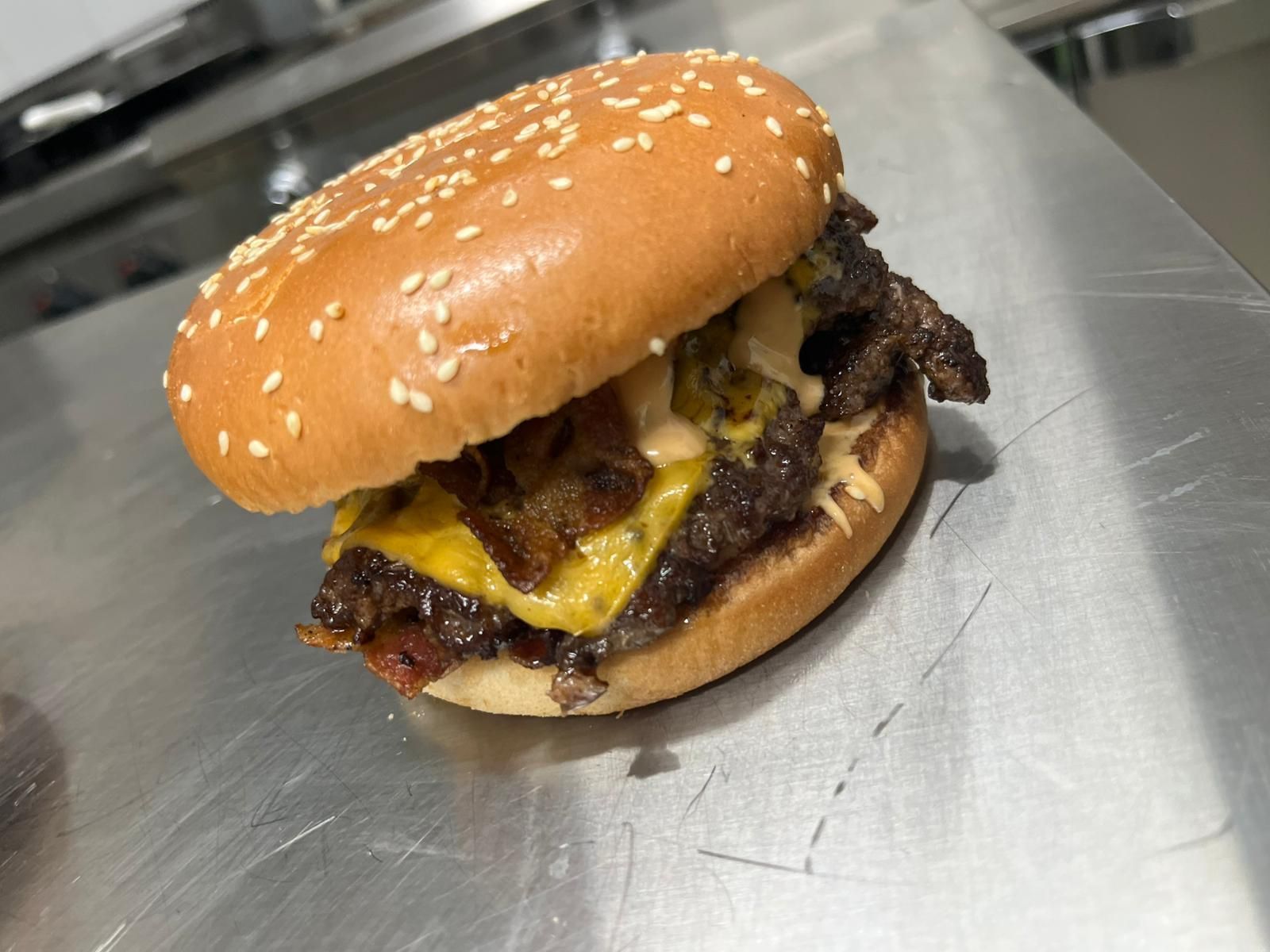 Cheeseburger con semi di sesamo, servito su una piastra in acciaio inox, con formaggio fuso e un tortino di carne scura.