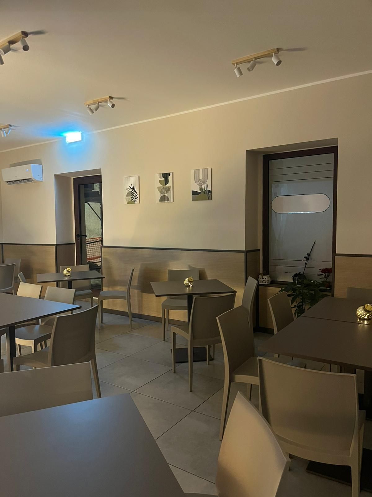 Interno vuoto di un caffè con tavoli e sedie, illuminazione calda, opere d'arte alle pareti e una porta aperta.