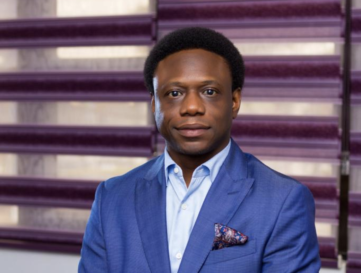 A man in a blue suit and white shirt is sitting in front of purple blinds.