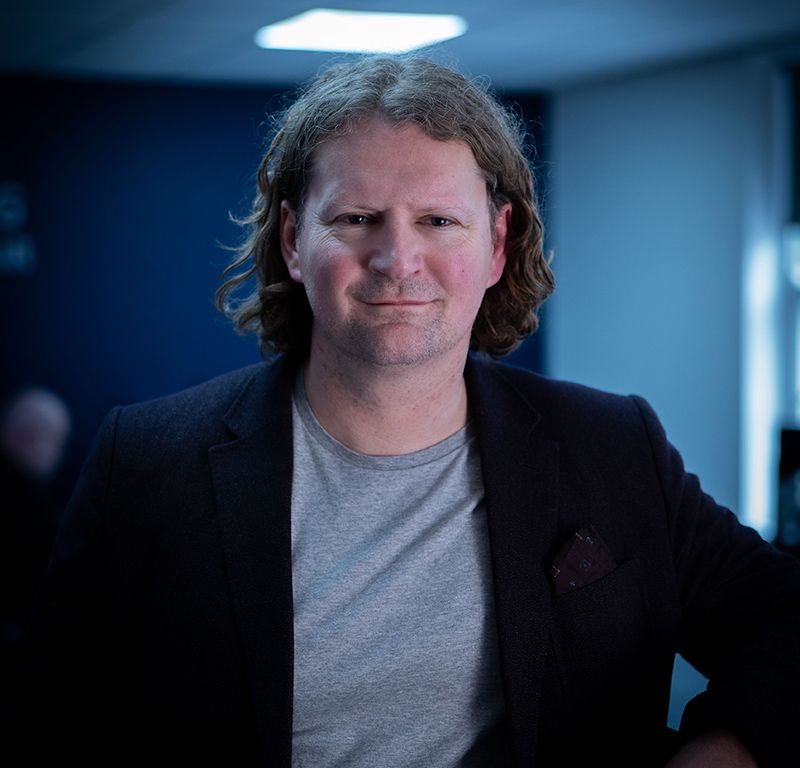 A man with long hair is wearing a black jacket and a grey shirt.