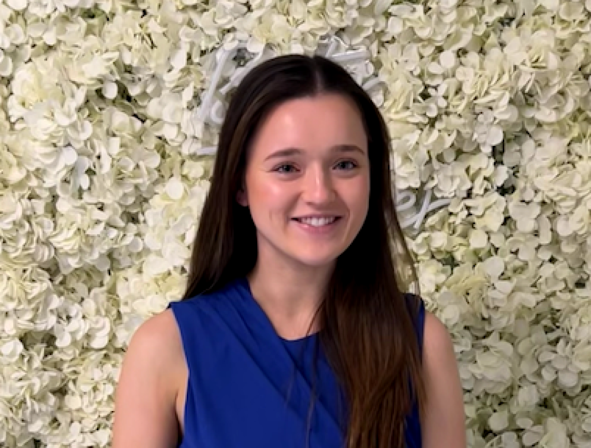 A woman in a blue shirt is smiling in front of a wall of white flowers.