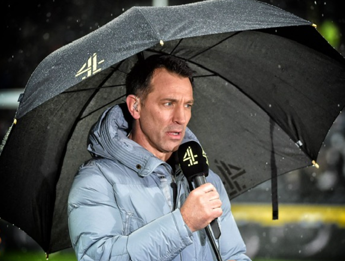 A man in a jacket is holding a microphone under an umbrella.