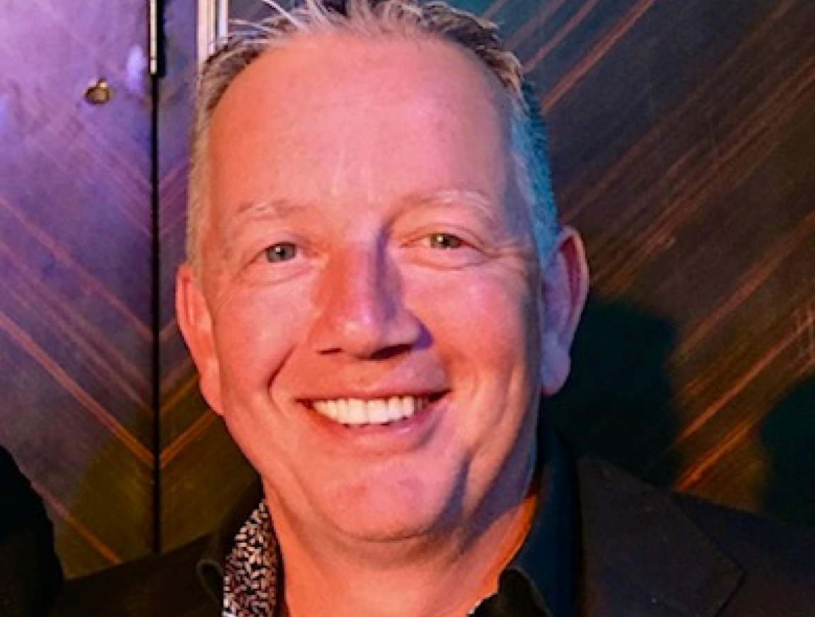 A man in a suit is smiling in front of a wooden wall.