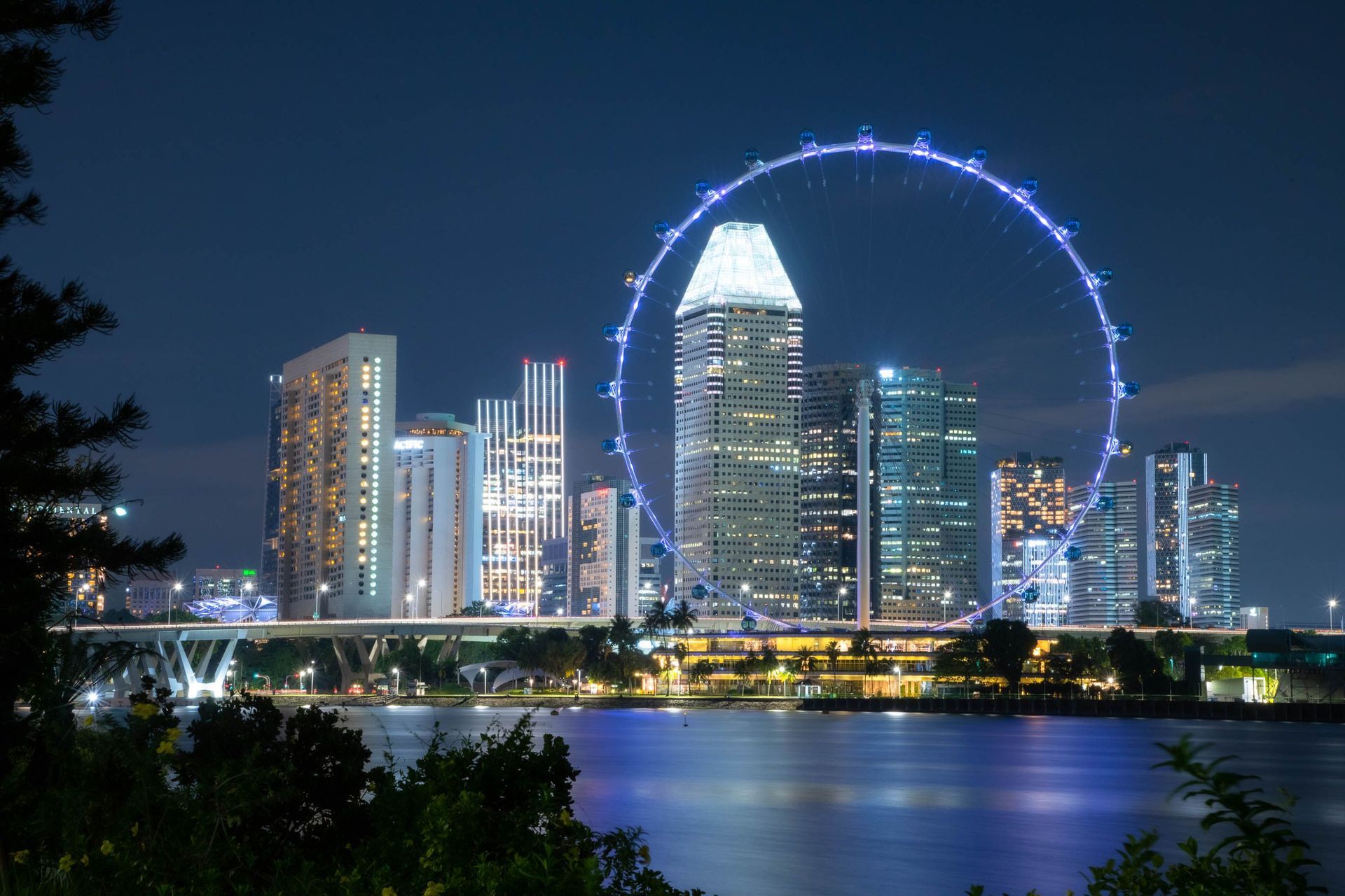 Night skyline with illuminated high-rises and a large lit Ferris wheel reflected on water