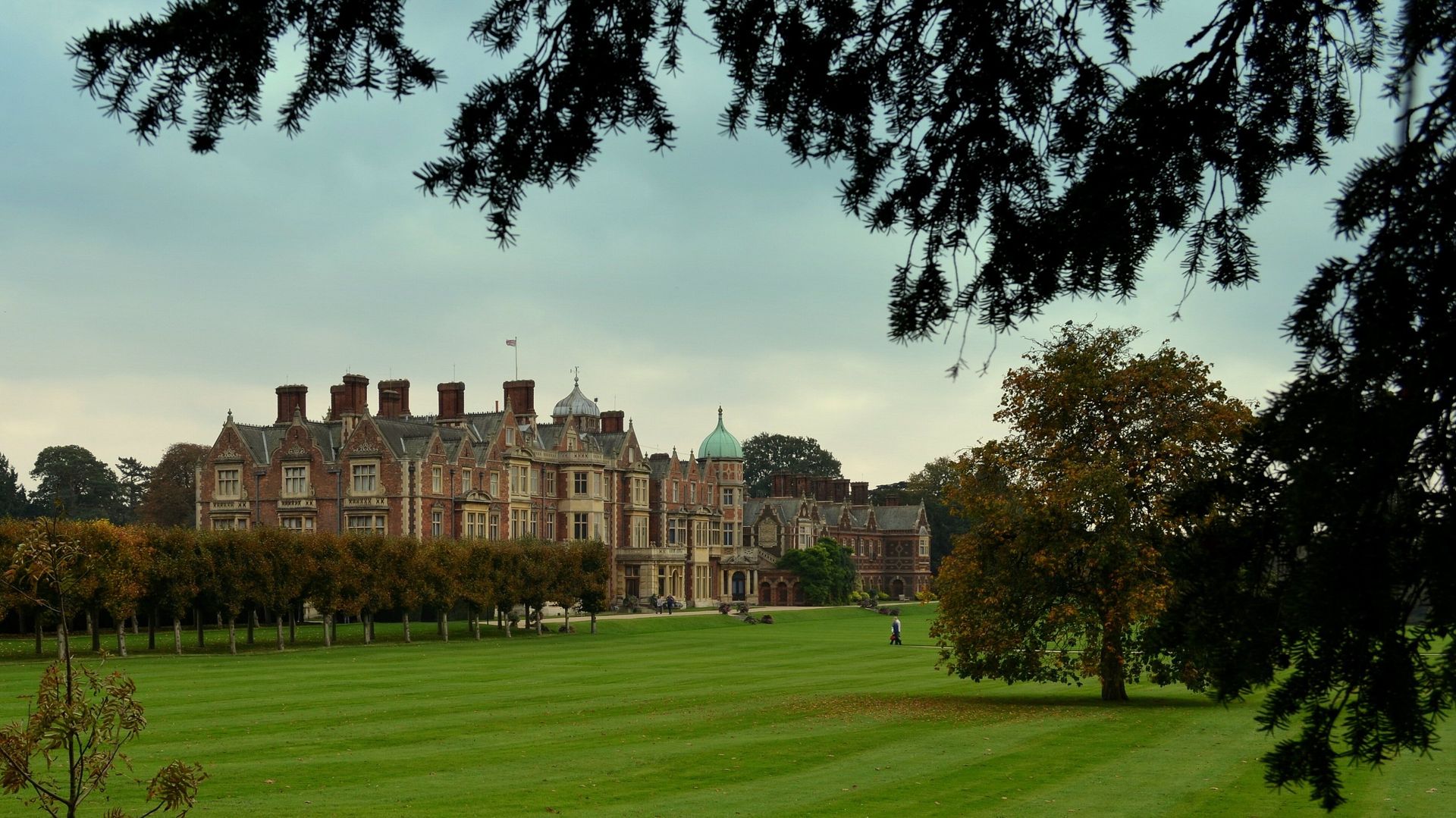 Large historic mansion with red-brick chimneys beside a wide green lawn, framed by tree branches.