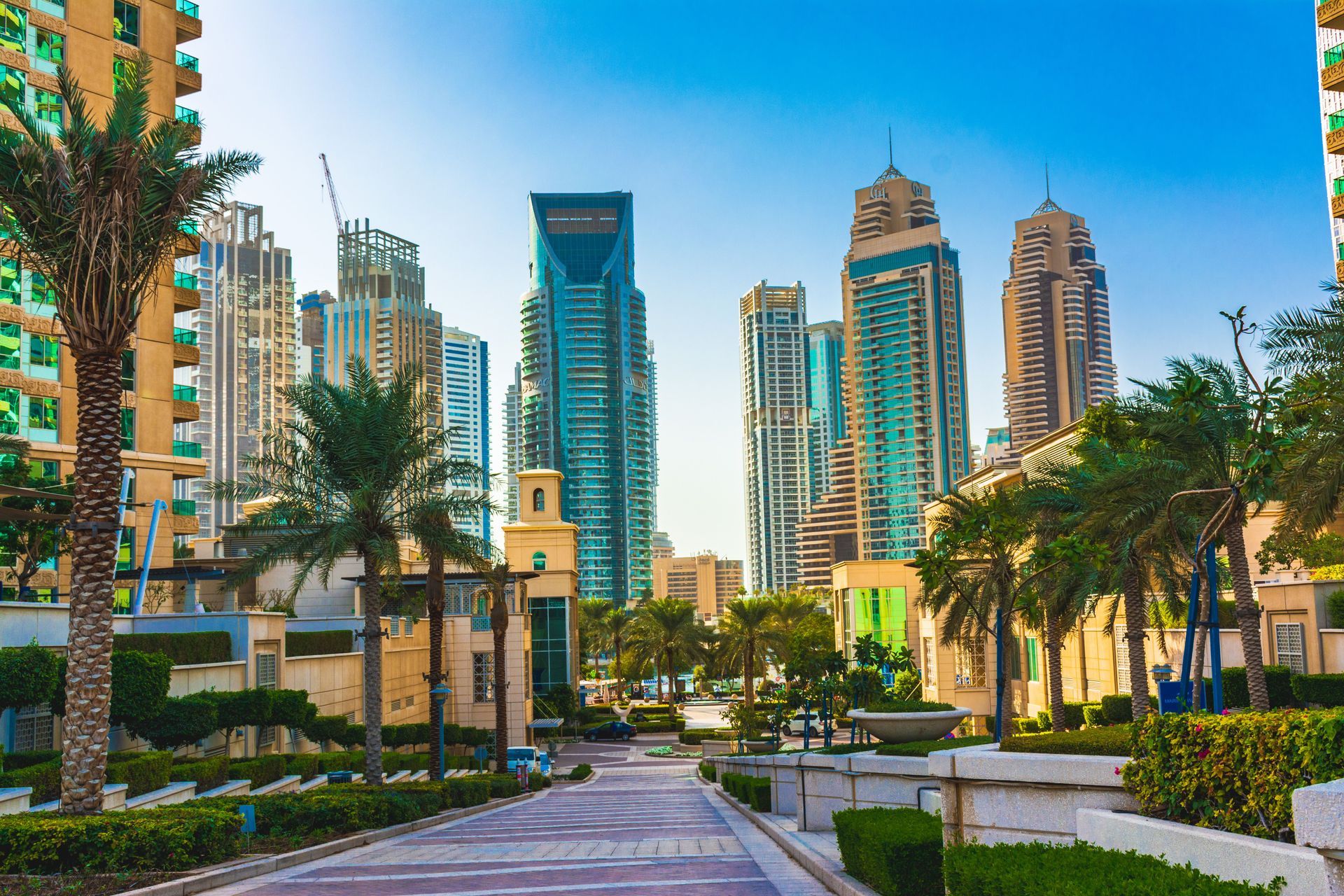 Modern city skyline with tall glass towers, palm trees, and a landscaped walkway under a blue sky