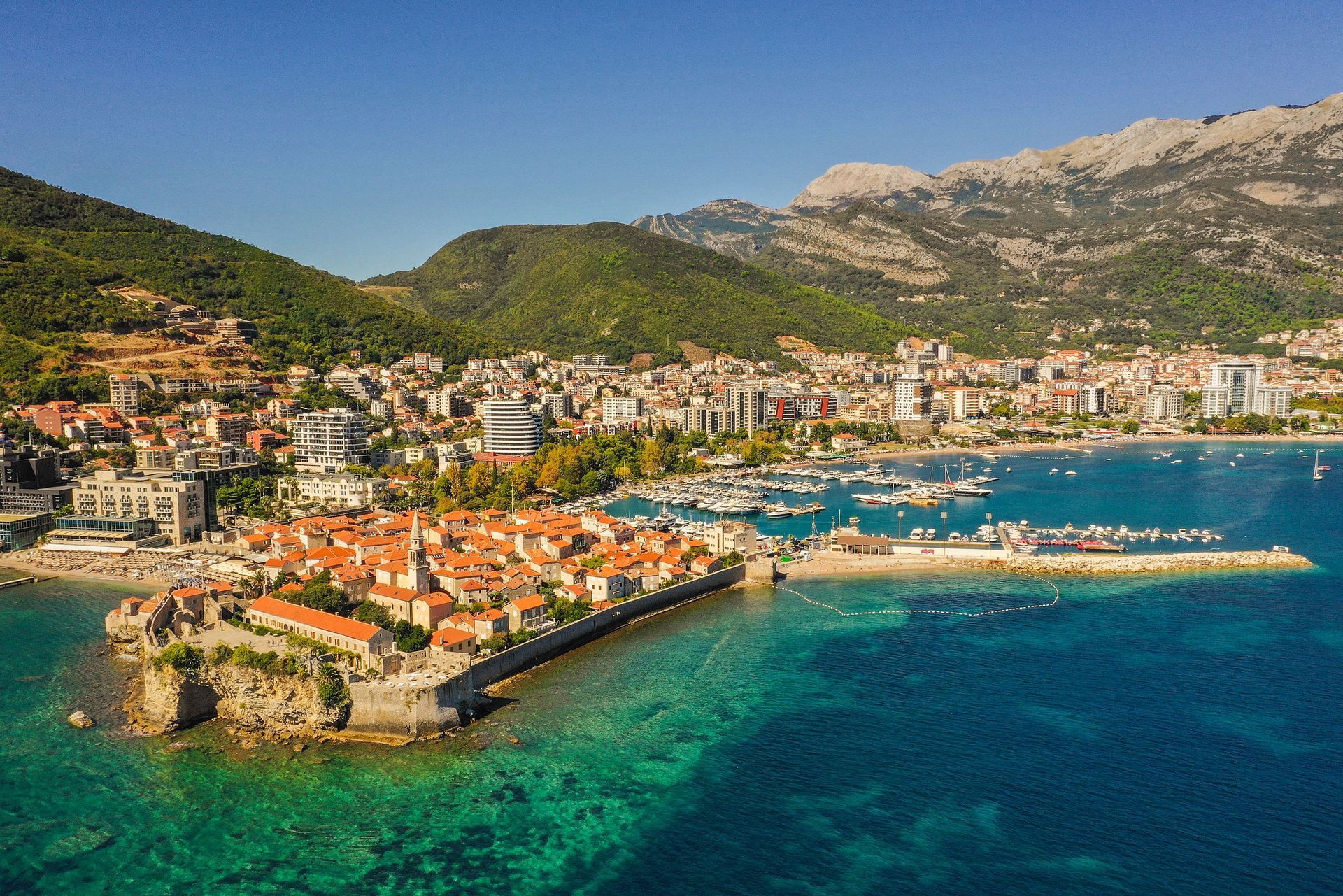 Aerial view of a coastal town with red roofs, turquoise bay, and mountains in the background