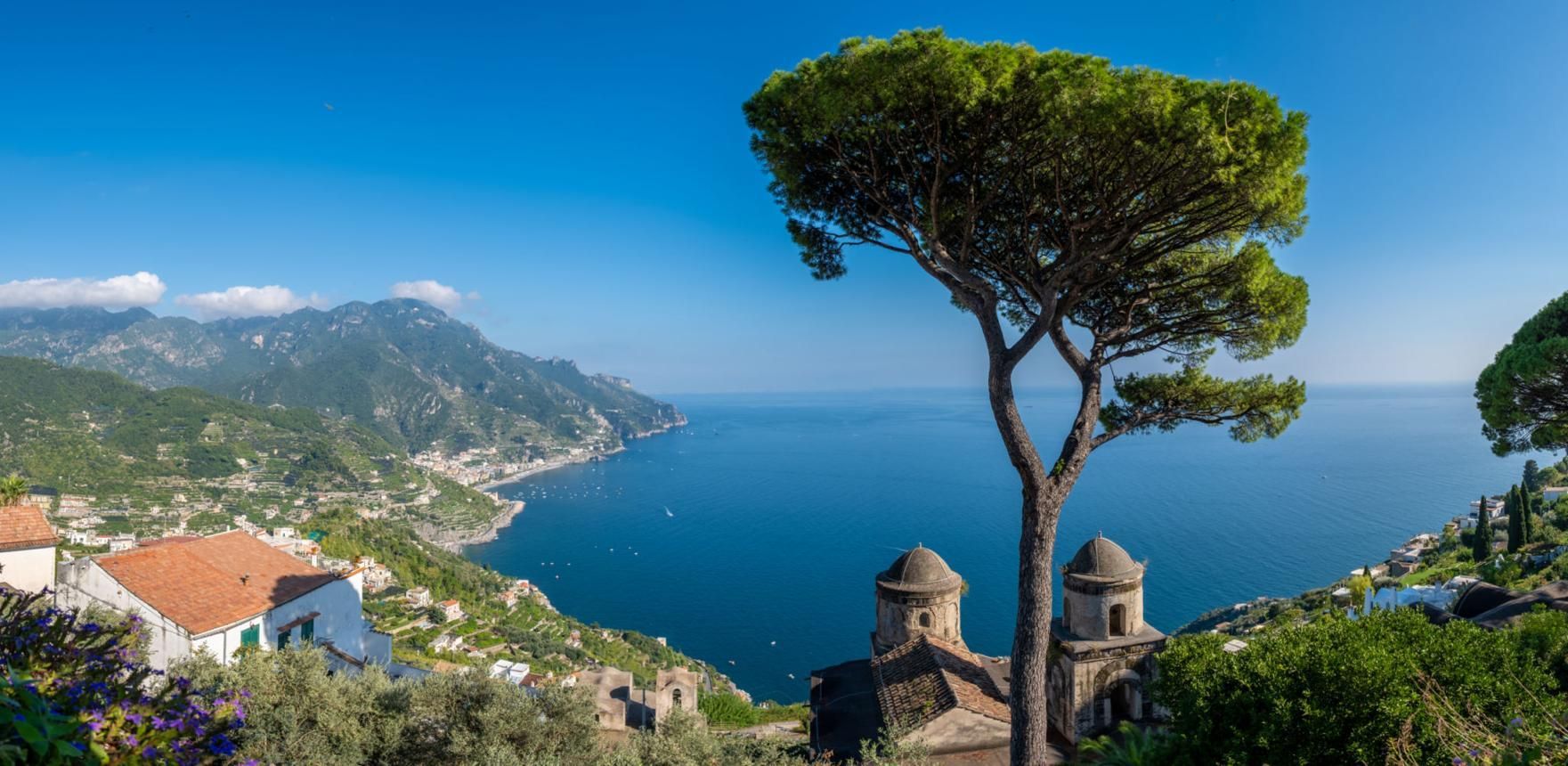 Coastal hillside village with sea view, umbrella pine, and distant mountains under a clear blue sky