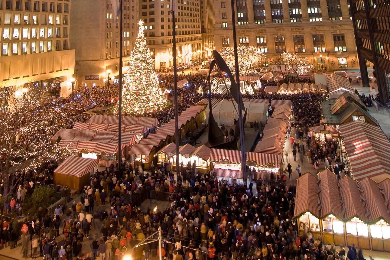 Crowded nighttime holiday market in a city square with lit stalls and a large Christmas tree