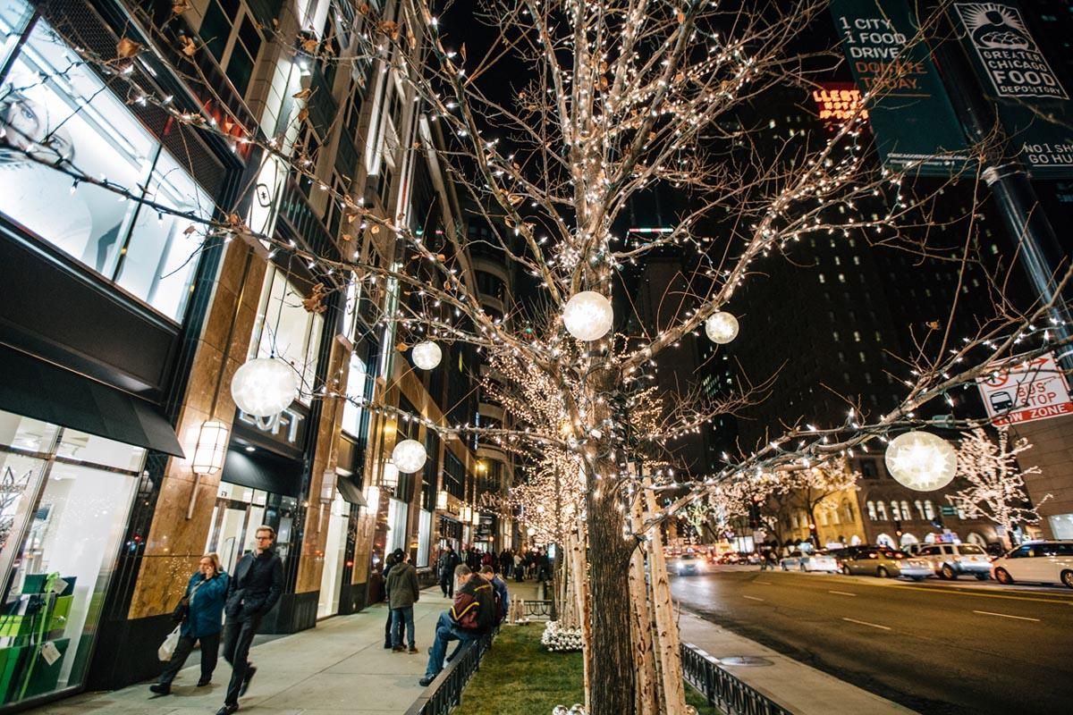 Night city sidewalk with lit trees, pedestrians, storefronts, and traffic passing by