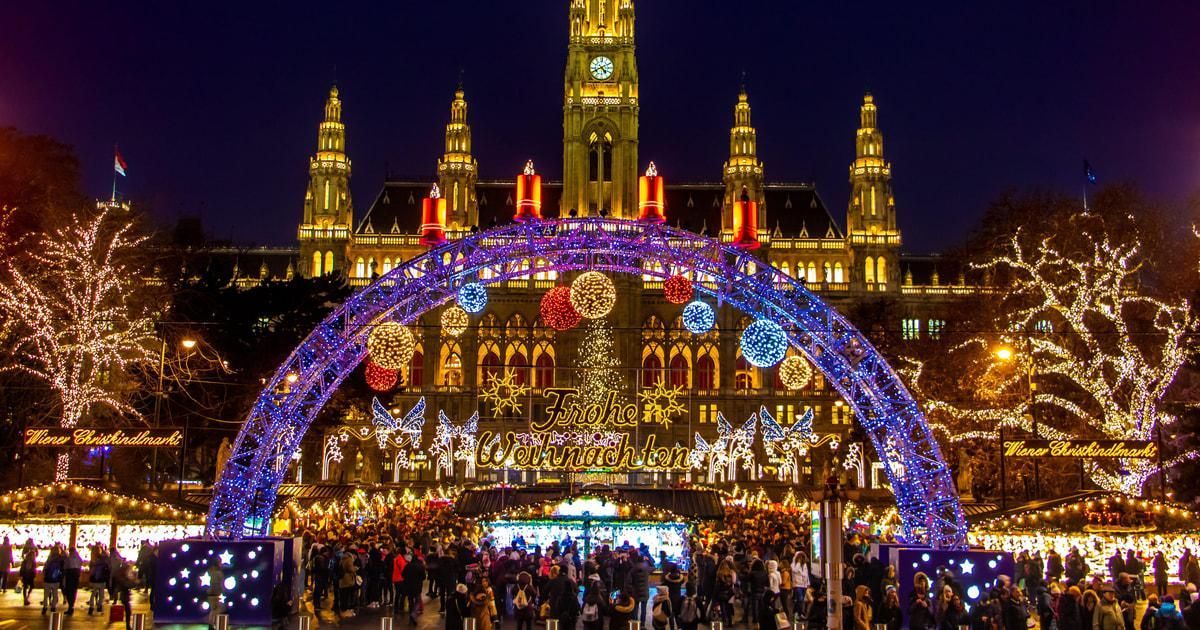 Lit-up holiday market with a glowing arch, festive crowd, and illuminated cathedral at night