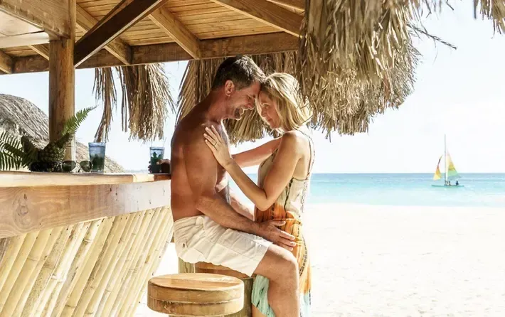 A couple embraces intimately at a beachside bar under a thatched roof, with a boat visible on the ocean in the background.