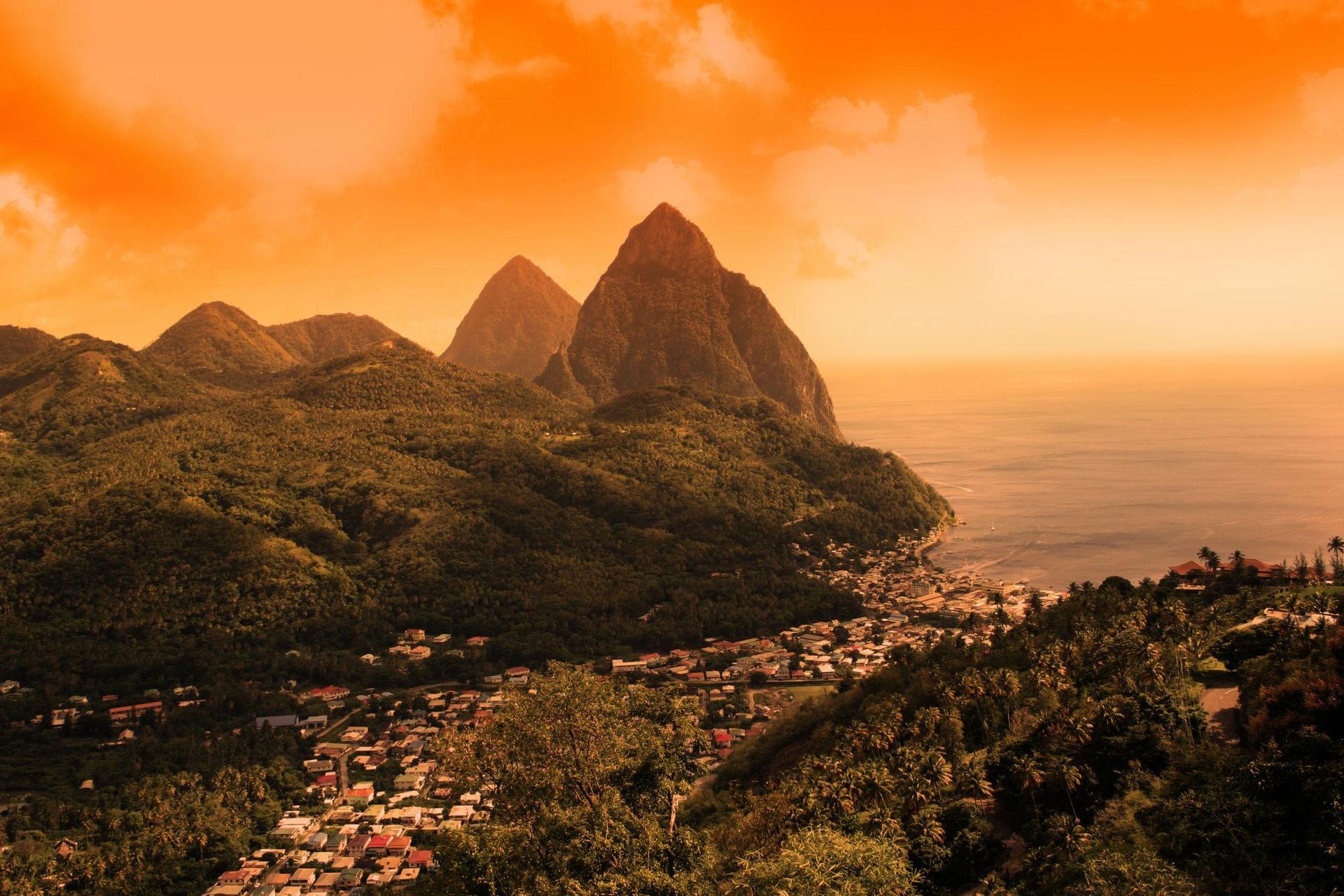 The Pitons mountains rise above a lush, green coastal town in St. Lucia under a vibrant orange sunset sky.