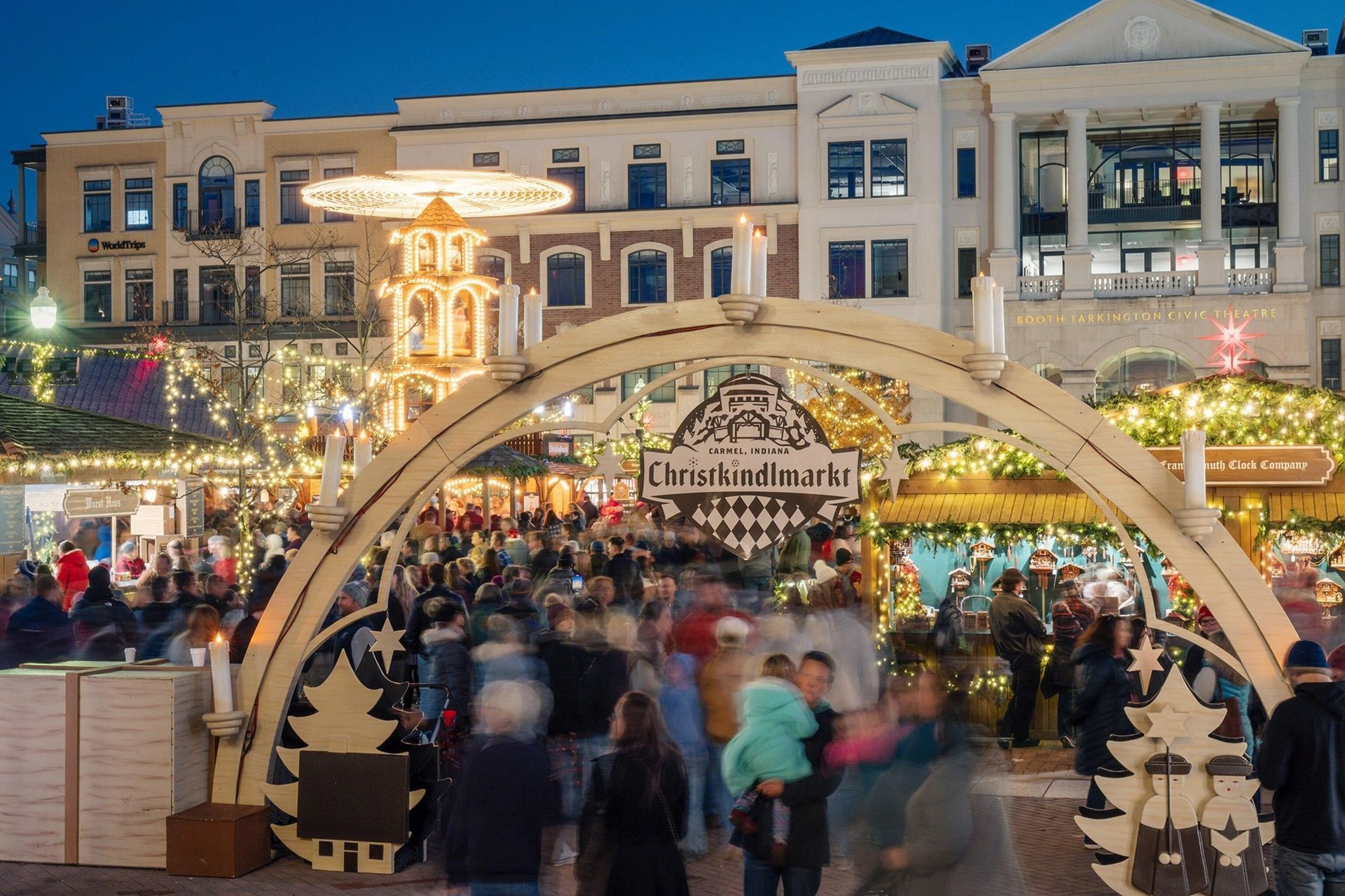 Crowded holiday market at dusk under a lit arch in front of a decorated building